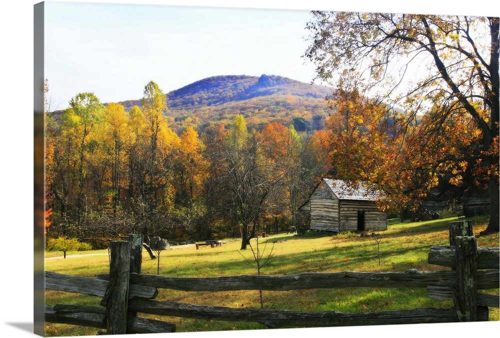 This is a landscape photograph of a log cabin in a meadow surrounded by autumn trees behind a primitive wood fence.