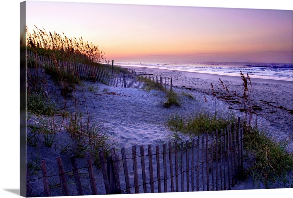 A landscape photograph of a dune covered in sea grass and fences fills the foreground of this beach at sunset.