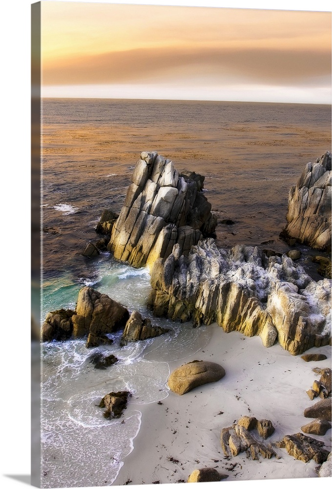 Vertical photo on canvas of tall rocks sticking out of the ocean by the shoreline with sun set warmed clouds in the sky.