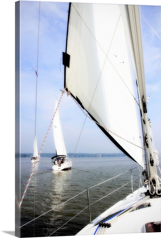 Big vertical photograph of a sail and the front of a sailboat, looking out toward the water where two other sailboats are ...