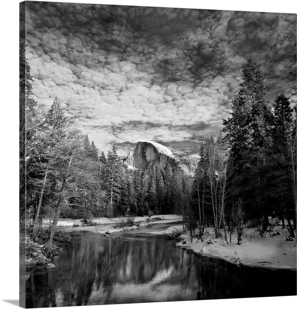 Square black and white landscape photograph of a body of water lined with snow and trees with a large rock formation in th...