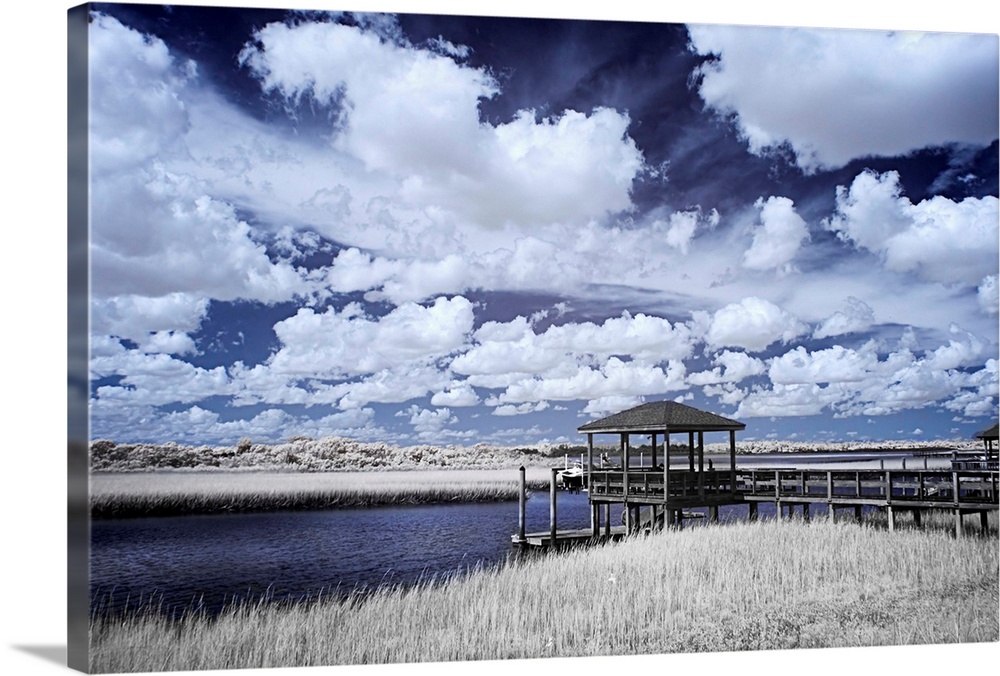 Cool toned photograph of a dock out to a river flowing through the marsh with beautiful, white, fluffy clouds in the sky.