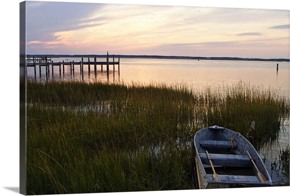 A boat sits in shallow water amidst tall grass at dusk by a pier as calm waters reflect the sun's rays.