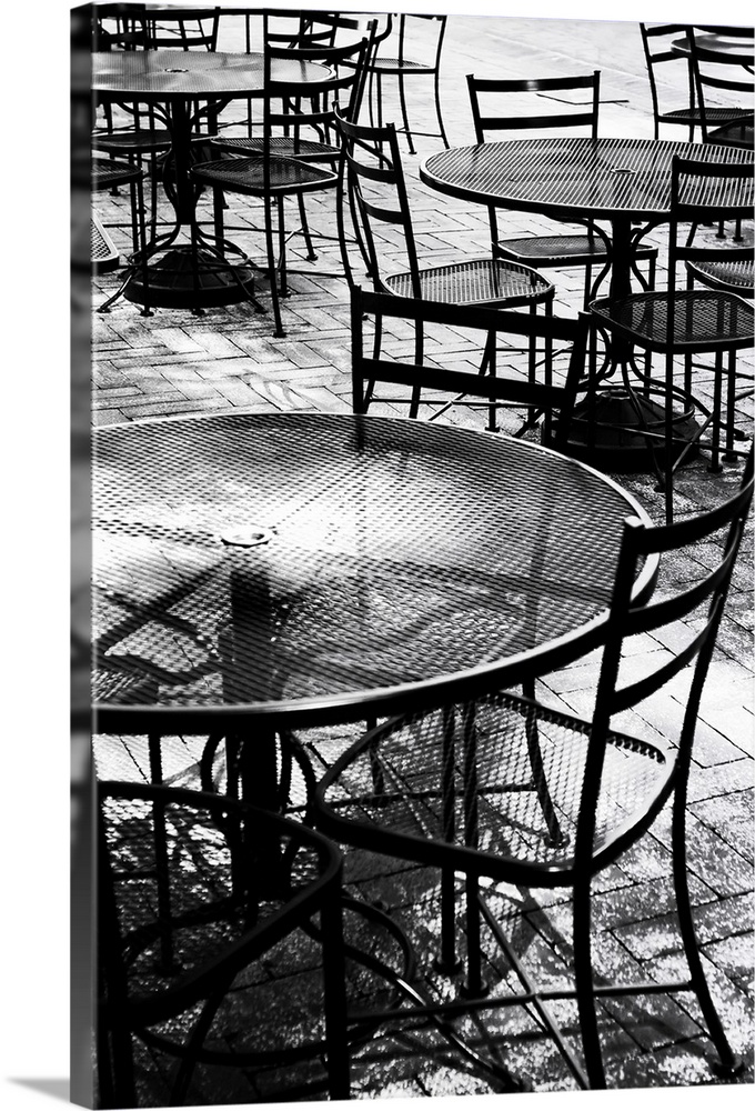 Black and white photograph taken of outdoor tables and chairs that sit on a brick sidewalk.
