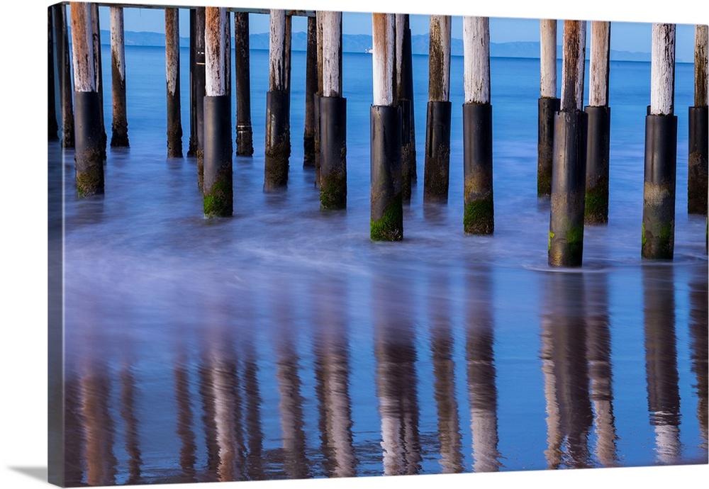 Photograph of the bottom of Ventura Pier pilings reflecting into the ocean water with mountains in the background.
