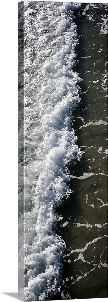 Tall, panoramic photograph of the white water from a crashing wave onto the shore.