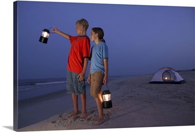 A boy and a girl stand with lanterns on the beach with a tent in the background