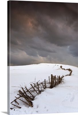 A Broken Fence Along A Snow Covered Field With Dark Clouds Overhead