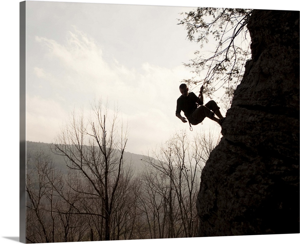 A rock climber is silhouetted against the sky as he rappels down the side of a cliff.