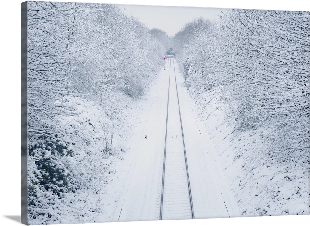 A view overlooking a snow covered railway line as it vanishes past a lone red signal in the distance, surrounded by snow c...