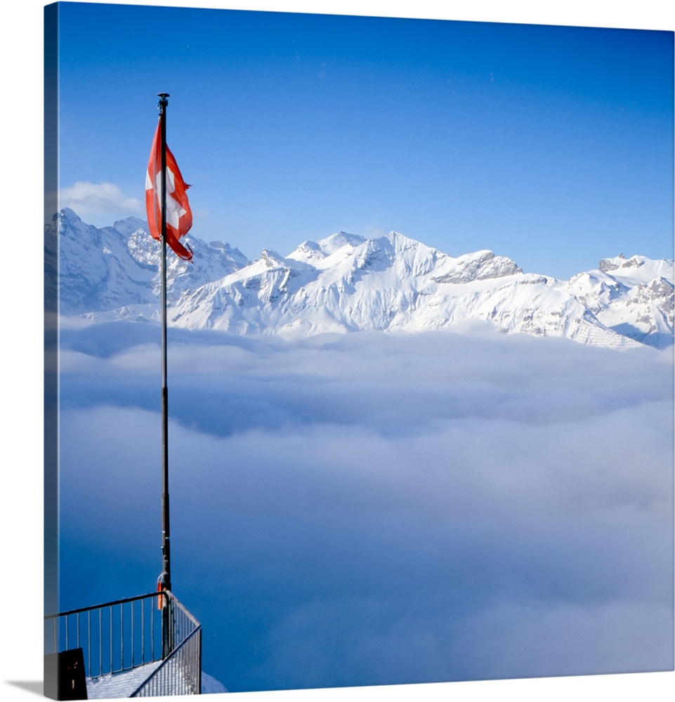Above-the-clouds panorama shot of Swiss Alps, Swiss flag can be seen in foreground.