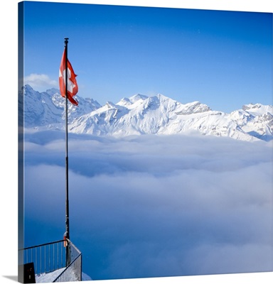 Above-the-clouds panorama shot of Swiss Alps, Swiss flag can be seen in foreground.