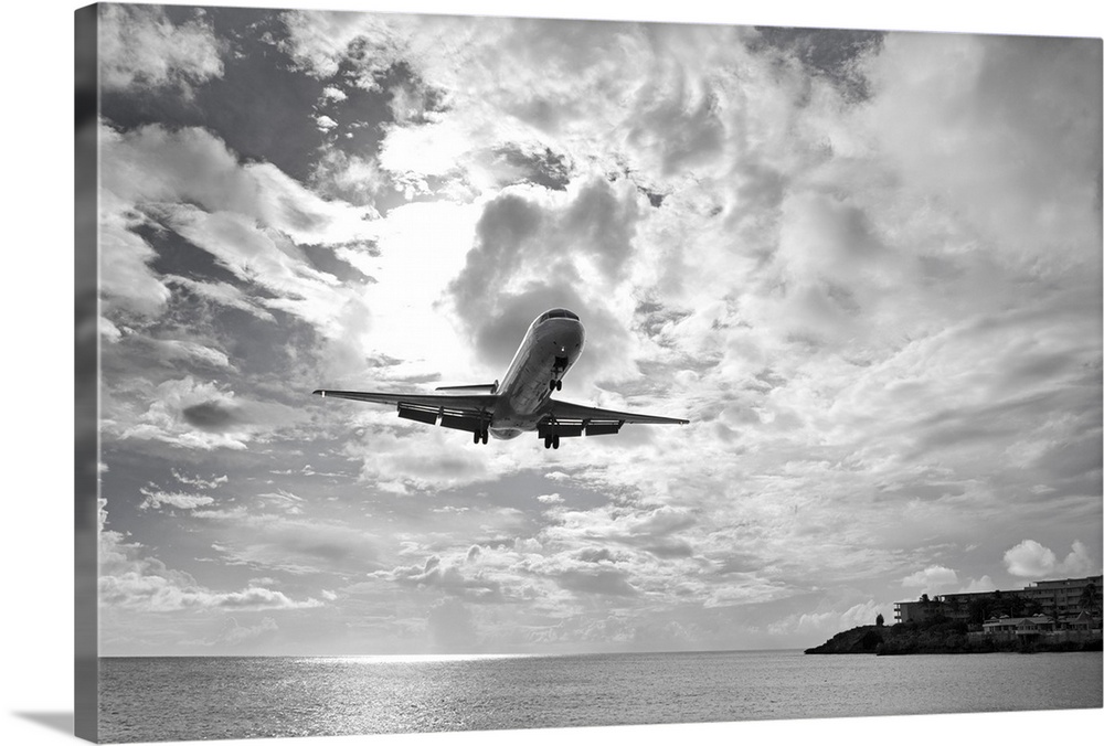 Black and white photograph taken of a commercial plane descending over ocean water about to land on an island.