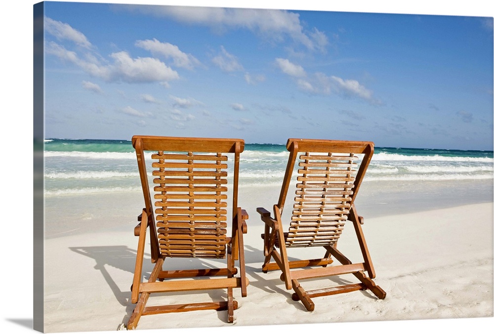 Large landscape photograph of two wooden lounge chairs sitting on the beach, beneath a blue sky.