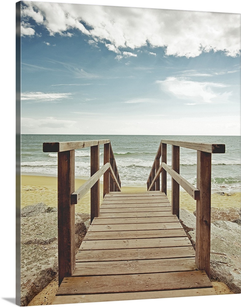 Photograph looking straight on of a wooden bridge spanning over large rocks that lead to a sandy beach and the ocean.