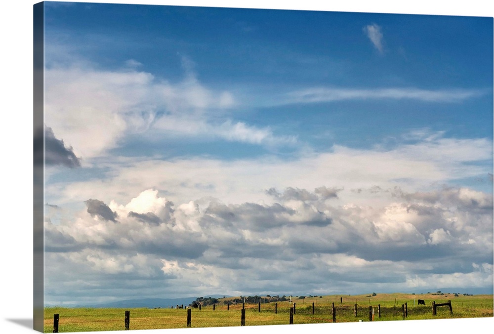Big blue sky with cumulus clouds fills most of image with slim view of grassland and fences of western ranch on lower quar...