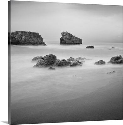 Black and White image of rocks in the ocean near a sandy beach.
