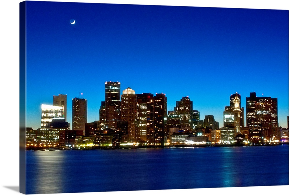 This big photograph is of the Boston skyline under a night sky and crescent moon. Buildings are lit and reflect in the water.