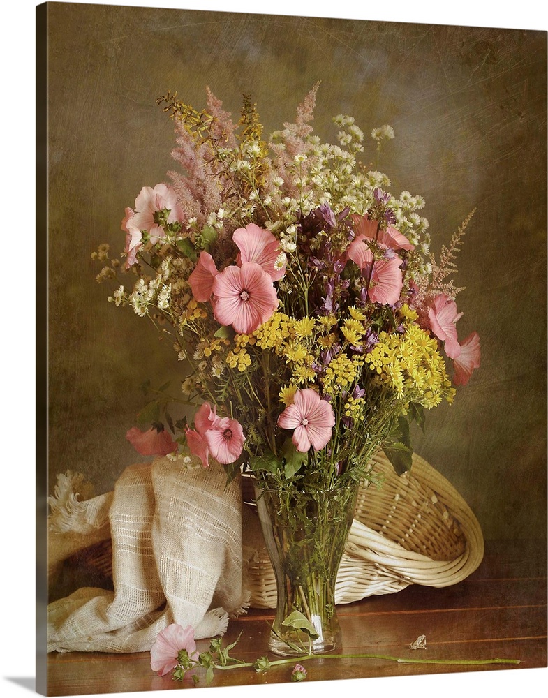 Lavatera and wild flowers in glass vase with basket with drapery in background.