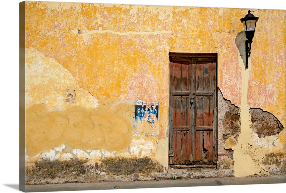 An old cracked wooden door on a bright yellow wall with crumbling stucco on a street in San Cristobal de las Casas, in Chi...