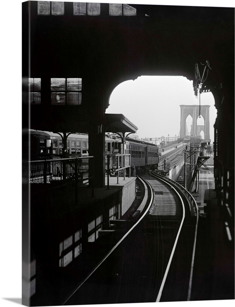 View of the Brooklyn Bridge from an elevated subway station on one shore. Photo shows a train beginning to cross the bridg...