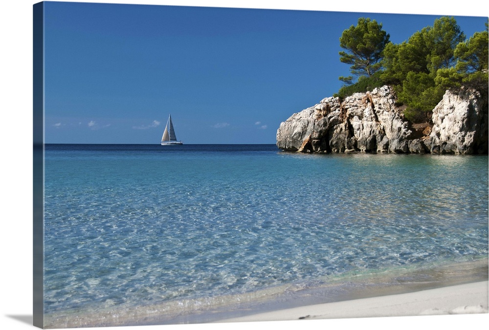 Photo of crystal clear blue water and white sand beach in Spain with a sailboat in the distance and a large cliff jutting ...