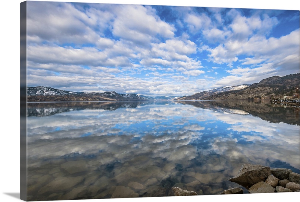 Late winter reflections of surrounding mountains and scattered clouds are mirrored in Okanagan lake.