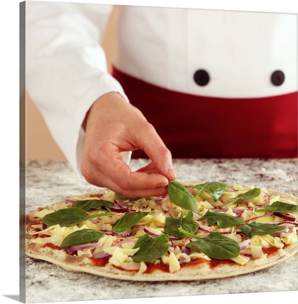 Close-up of a chef preparing a pizza. Hands placing spinach on pizza topped with cheese and tomato sauce. Granite workbench.