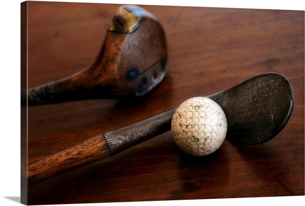 Horizontal photograph on a large canvas of two antique golf clubs and a ball, lying on a wooden table.