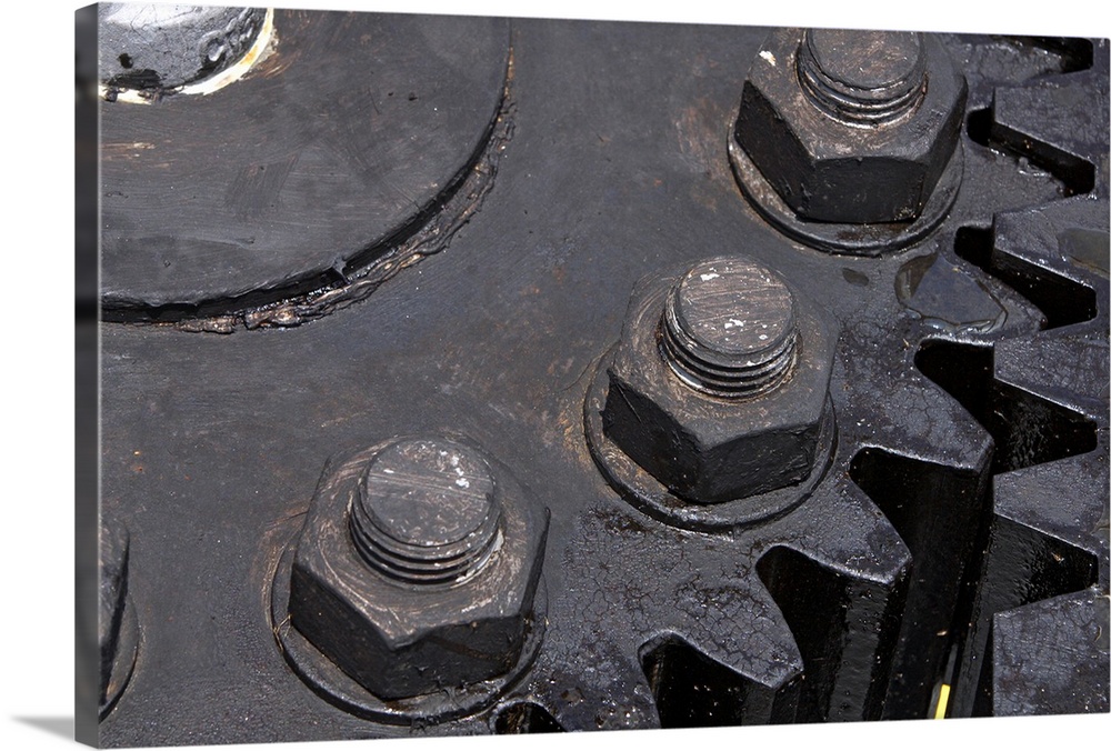 Oversized, horizontal, close up photograph of the teeth meeting on two large metal gears. Large bolts can be seen on the e...