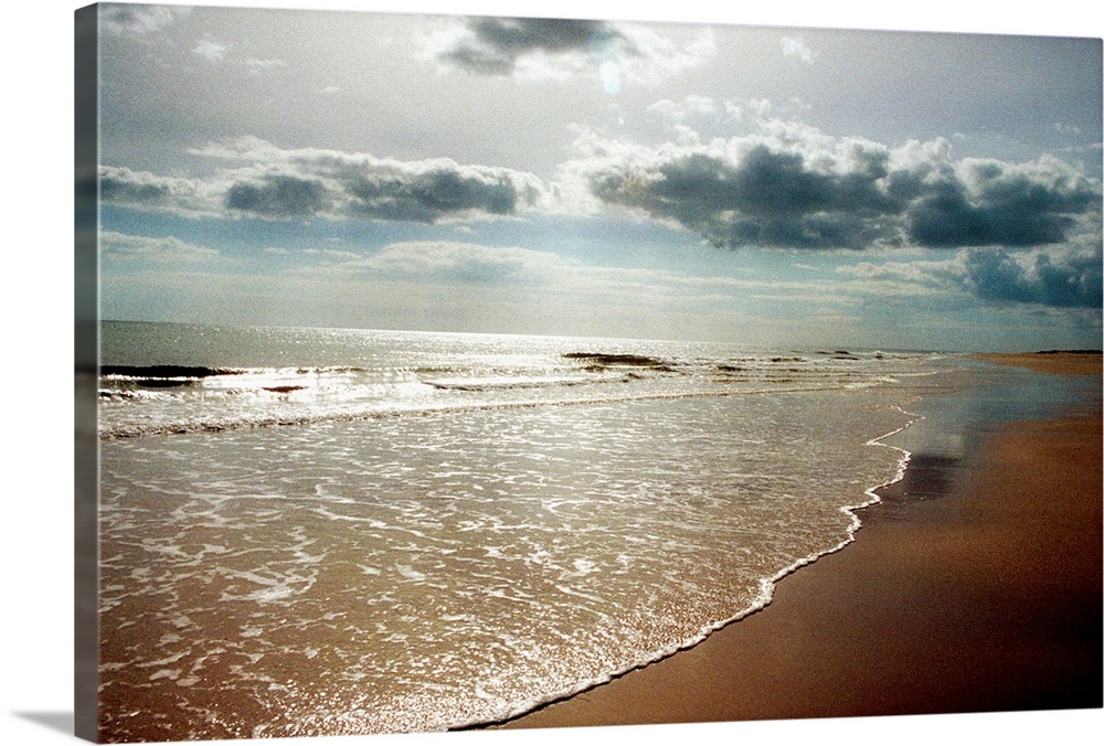 Sunlight shining down through puffy clouds onto a sandy beach and shallow ocean waves flowing onto the shore.
