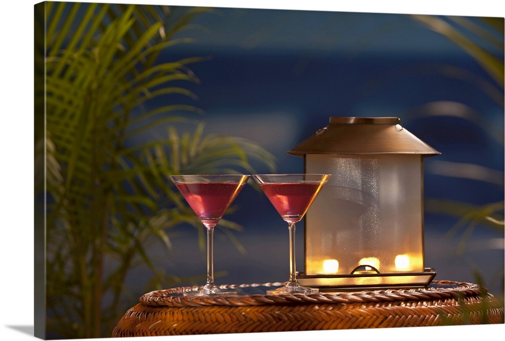 Photograph of two martini glasses with a lantern sitting on a table with palm trees and ocean in the distance.