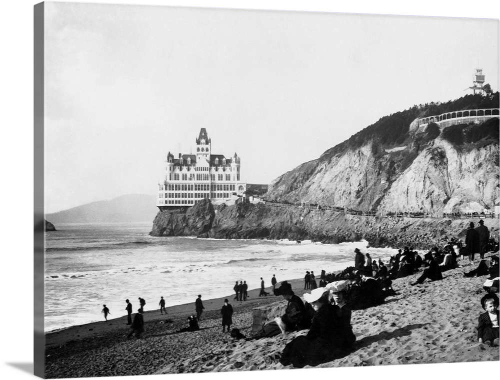 A crowd of people on the beach below the Cliff House enjoy the sun, the surf, and a view of Seal Rocks. The Gothic Revival...