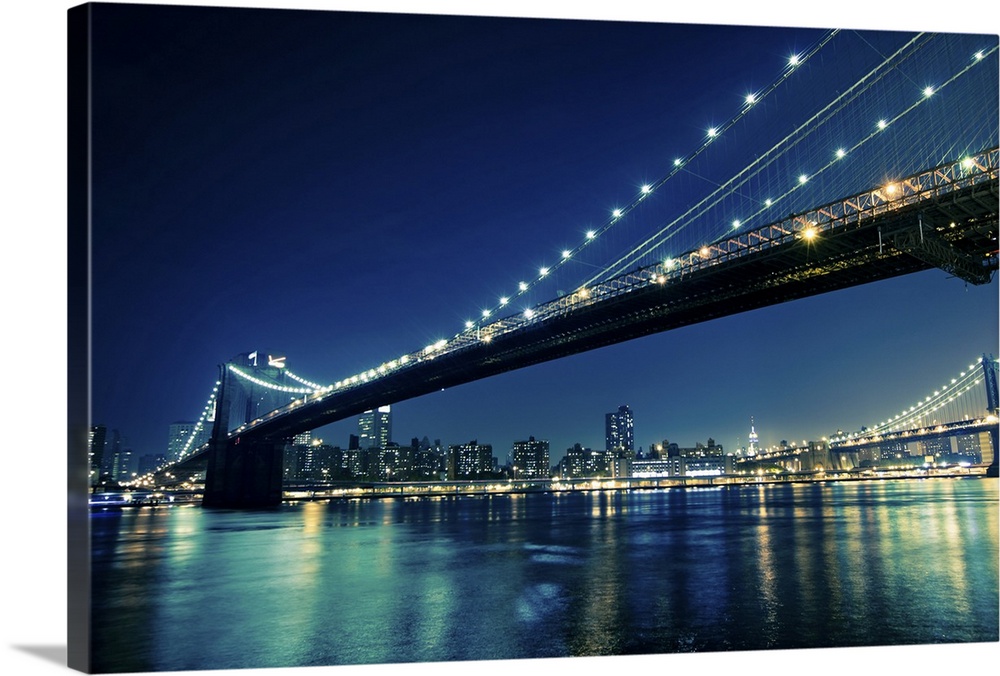 The city of New York and bridges that stretch across the river are illuminated under a night sky and reflect back into the...