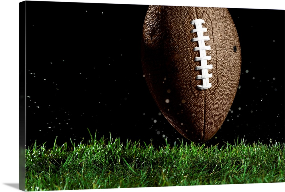Giant landscape photograph of a football in a vertical position as it moves above the wet grass with specks of water flyin...
