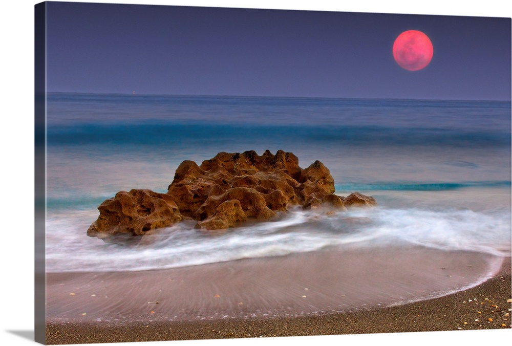 A unique rock formation sits in the ocean water which is photographed under a deep red moon.