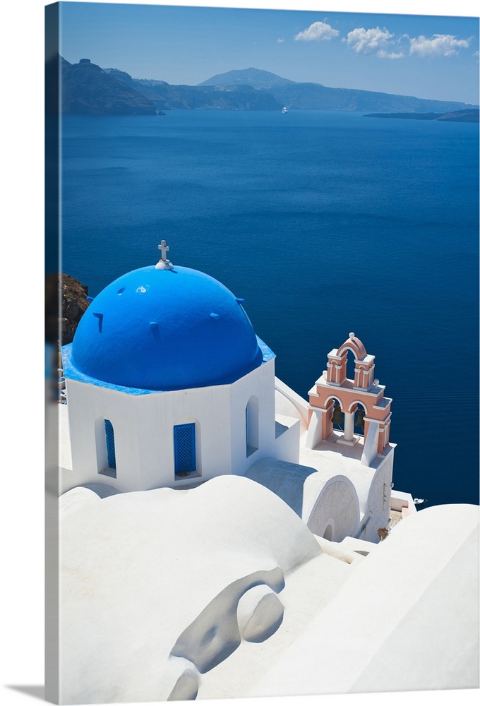 Oversized, vertical, aerial photograph of the top of a church, overlooking blue waters over the coast of Oia, Santorini, G...