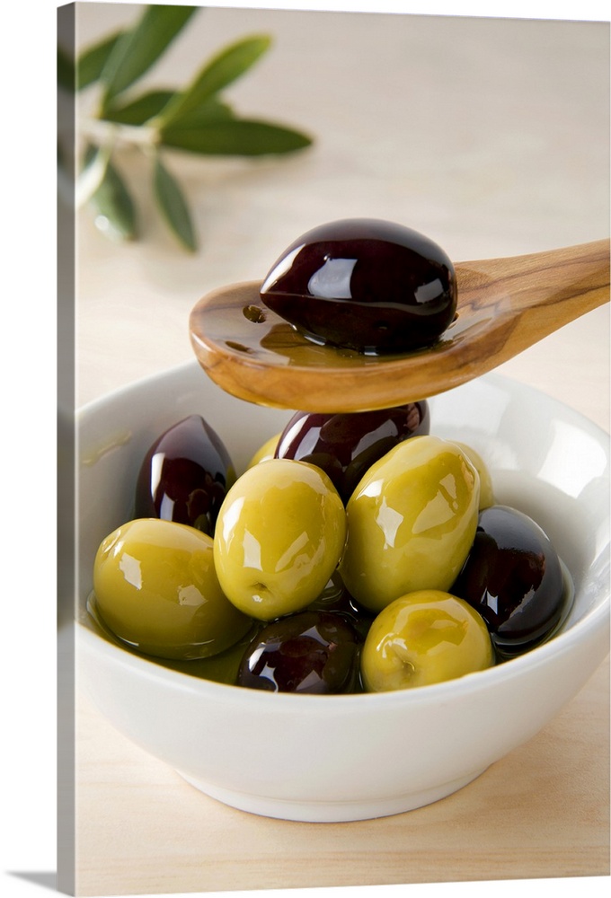 Green and black olives covered in olive oil in a bowl and on a wooden spoon, close-up photograph.