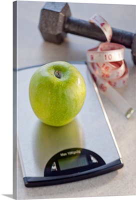 Green apple on weight scale, tape measure and exercise weight in background