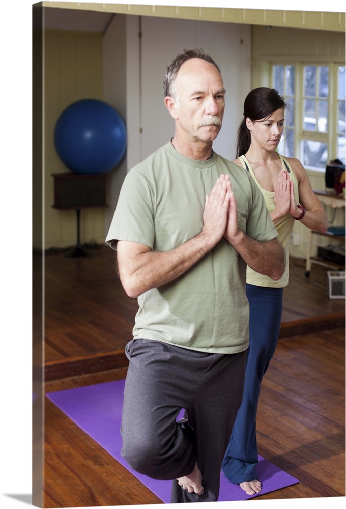 An older man in front of a younger woman practices a yoga position in a small studio.