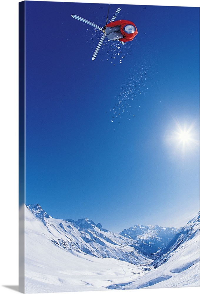 Vertical photo of a man crossing his skis on as he flies through the air as the sun shines down on the snow covered mounta...