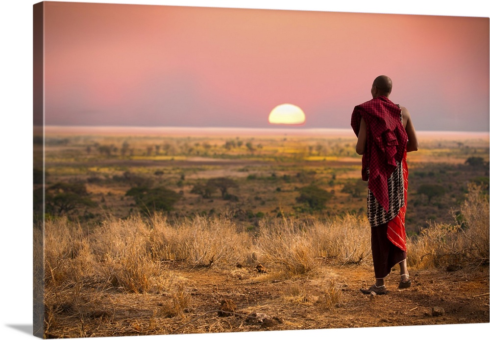 Masai man, wearing traditional blankets, overlooks Serengeti in Tanzania as the colorful sunset fills the sky.