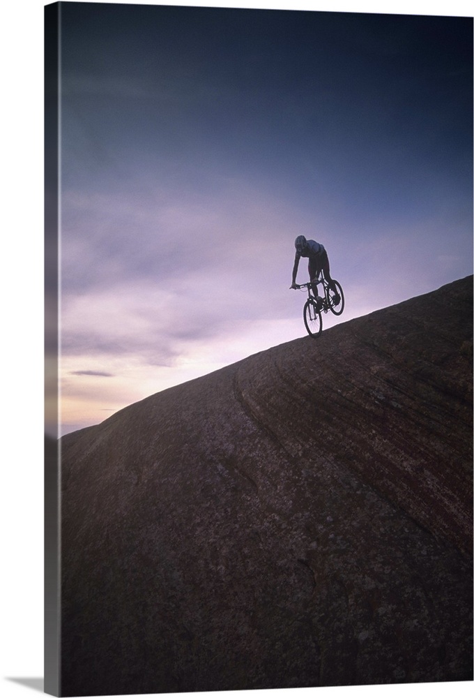 Mountain biker on desert rock formation silhouetted against sky at sunset