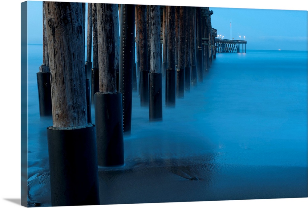 Large print of tall wooden pier pillars holding up a pier leading into the ocean from the shore.