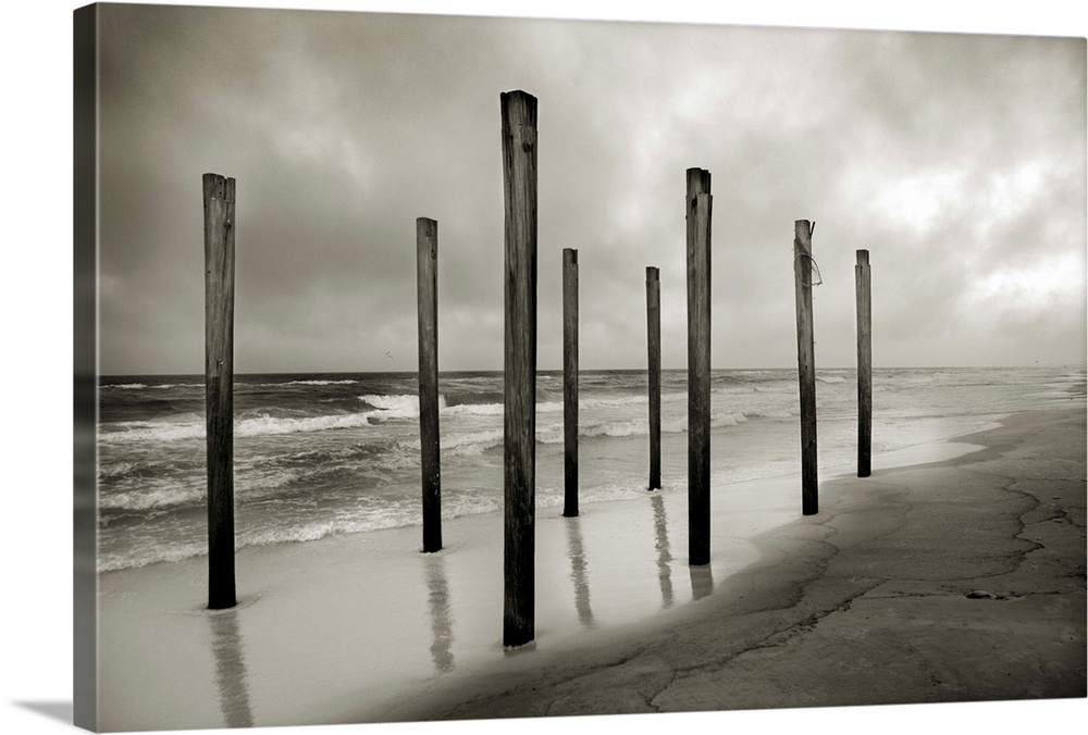 A sepia toned shot of old beach posts at the shoreline with clouds and ocean in the background.