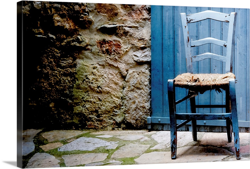 Front capture of old blue wooden chair with damaged caned seat, at doorstep of house with blue wooden door and stone pavem...