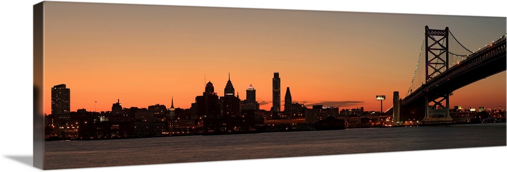 Long horizontal image of the silohuette of the skyline of Philadelphia along the water front with a bridge on the right le...
