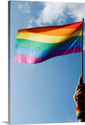 Person's hand holding rainbow-colored flag waving in the sky