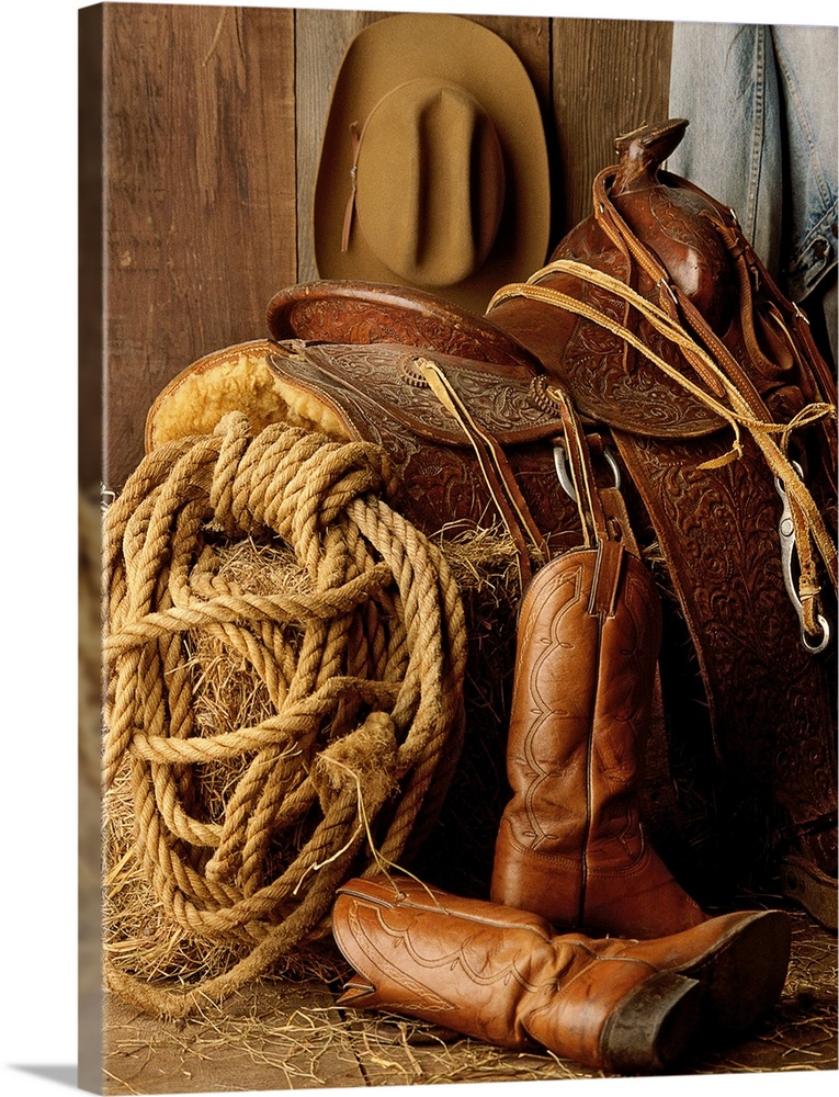 Staged photo of a horse saddle on a bale of hay surrounded by other Cowboy necessities such as a lasso, cowboy hat, and bo...