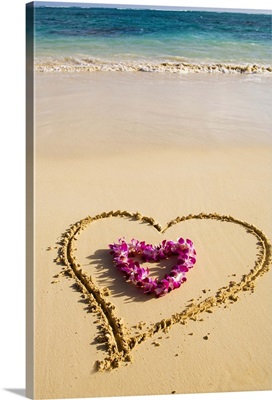 Picture of a heart drawn in the sand, orchid lei shaped like a heart inside of drawing